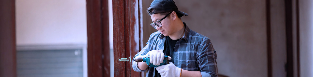 Person drills into a wall frame while wearing gloves and a backward cap.