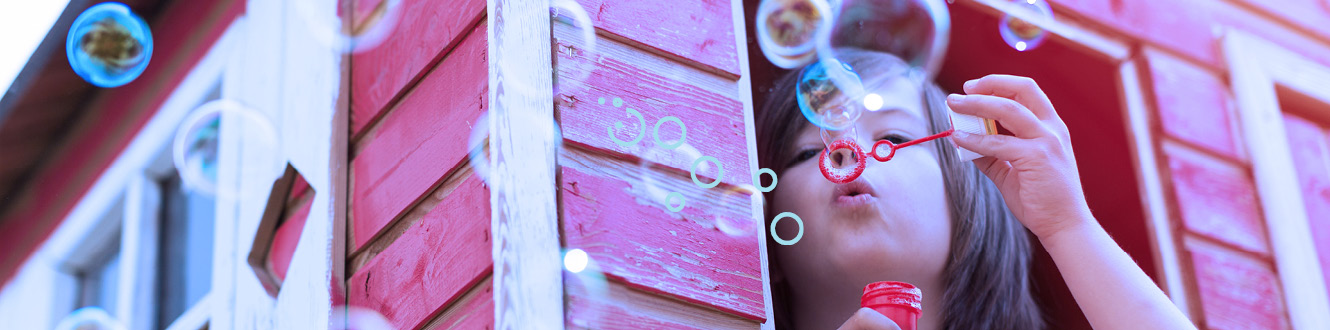 A young child in a play house blowing bubbles out the window. 